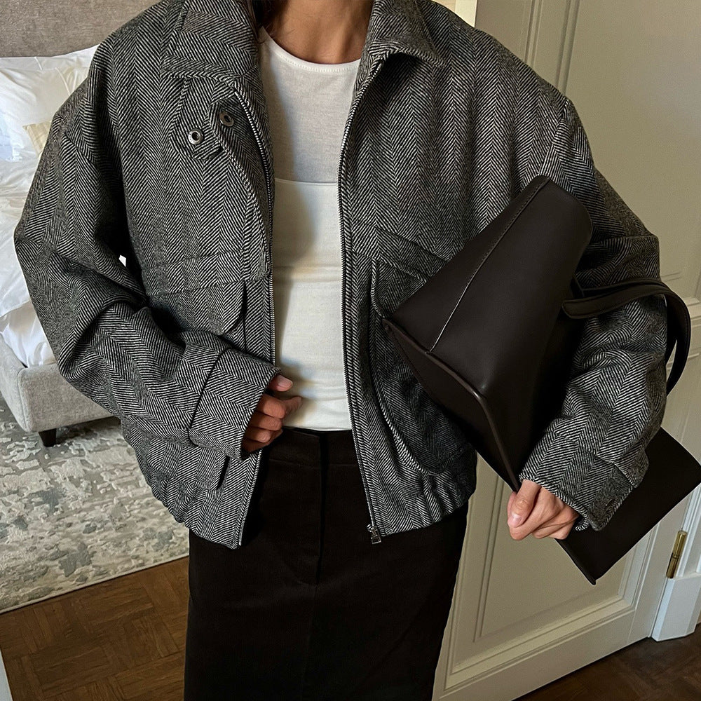 Person wearing a gray jacket holding a black bag in a room with a bed and carpet.